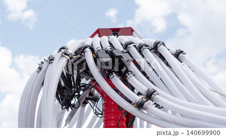 Close-up view of a red industrial machine topped with many white flexible hoses, black connectors, and bundled cables rising into a bright blue sky with soft clouds, creating a Close-up view of a red industrial machine topped with many white flexible hoses, black connectors, and bundled cables rising into a bright blue sky with soft clouds, creating a 140699900