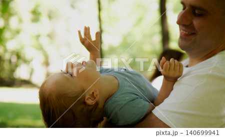 Father cradling young toddler on his arms while child laughs and reaches upward in a sunlit leafy park setting; warm natural light, shallow depth of field Father cradling young toddler on his arms while child laughs and reaches upward in a sunlit leafy park setting; warm natural light, shallow depth of field 140699941