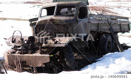Weathered midcentury flatbed truck with corroded metal cab and wooden stake bed sitting partially collapsed in melting snow on rural field; broken windshield Weathered midcentury flatbed truck with corroded metal cab and wooden stake bed sitting partially collapsed in melting snow on rural field; broken windshield 140699981