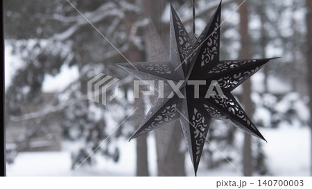 Black paper star ornament suspended near glass window overlooking snow-covered evergreen trees and soft winter landscape Black paper star ornament suspended near glass window overlooking snow-covered evergreen trees and soft winter landscape 140700003