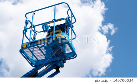 Blue elevated work platform with safety railings and metal mesh basket raised high against a bright blue sky with white clouds, viewed from below Blue elevated work platform with safety railings and metal mesh basket raised high against a bright blue sky with white clouds, viewed from below 140700034