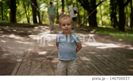 Small boy stands centered on a paved tree-lined park walkway, hands behind back and a gentle smile Small boy stands centered on a paved tree-lined park walkway, hands behind back and a gentle smile 140700037