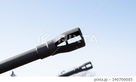 Low-angle closeup shows matte black artillery barrel muzzle pointing upward against pale blue sky, with blurred secondary barrel and faint treeline in Low-angle closeup shows matte black artillery barrel muzzle pointing upward against pale blue sky, with blurred secondary barrel and faint treeline in 140700085