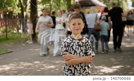 Smiling young boy stands in foreground with arms crossed wearing a patterned T-shirt while blurred families and children walk along a tree-lined paved park Smiling young boy stands in foreground with arms crossed wearing a patterned T-shirt while blurred families and children walk along a tree-lined paved park 140700088