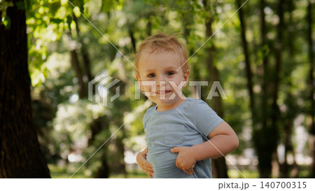 A cheerful young boy stands outdoors in a sunlit park surrounded by trees, wearing a light blue shirt A cheerful young boy stands outdoors in a sunlit park surrounded by trees, wearing a light blue shirt 140700315