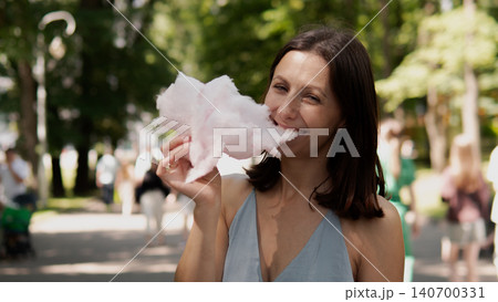 A smiling woman in a blue sleeveless dress enjoys cotton candy at a sunny park A smiling woman in a blue sleeveless dress enjoys cotton candy at a sunny park 140700331