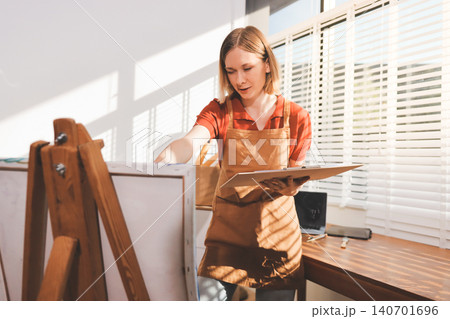 Picture of serious concentrated young Caucasian female artist sitting at desk with painting accessories, holding tube of oil paint, mixing colors on palette; unfinished painting on canvas near her Picture of serious concentrated young Caucasian female artist sitting at desk with painting accessories, holding tube of oil paint, mixing colors on palette; unfinished painting on canvas near her 140701696