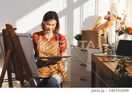Picture of serious concentrated young Caucasian female artist sitting at desk with painting accessories, holding tube of oil paint, mixing colors on palette; unfinished painting on canvas near her 140701699