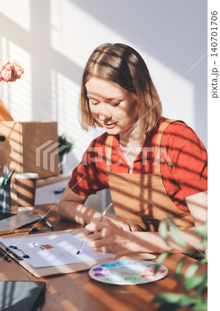 Picture of serious concentrated young Caucasian female artist sitting at desk with painting accessories, holding tube of oil paint, mixing colors on palette; unfinished painting on canvas near her 140701706
