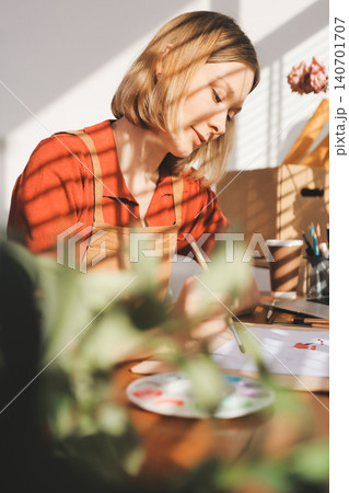 Picture of serious concentrated young Caucasian female artist sitting at desk with painting accessories, holding tube of oil paint, mixing colors on palette; unfinished painting on canvas near her 140701707