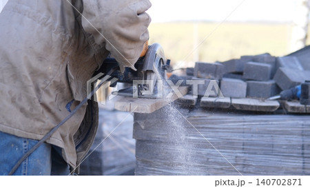 Worker Cutting Paving Blocks With Construction Grinder, Sparks And Dust Rising As Mason Trims Edges On Stacked Pallet, Power Tool And Protective Gloves In Industrial Outdoor Setting. Worker Cutting Paving Blocks With Construction Grinder, Sparks And Dust Rising As Mason Trims Edges On Stacked Pallet, Power Tool And Protective Gloves In Industrial Outdoor Setting. 140702871