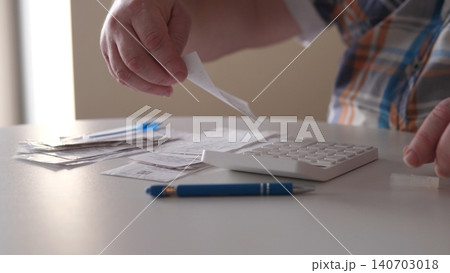 A man is sorting through receipts at a table while using a calculator. He holds a receipt in one hand and has papers spread out around him. A pen sits nearby. A man is sorting through receipts at a table while using a calculator. He holds a receipt in one hand and has papers spread out around him. A pen sits nearby. 140703018
