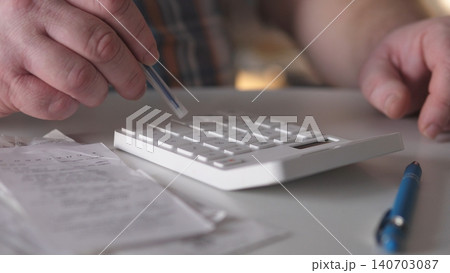 A person presses buttons on a calculator while handling receipts at a desk. Papers are spread out around, and a blue pen rests on the surface nearby as afternoon light shines in. 140703087