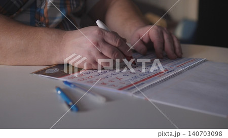 A person is using a red pen to write on a calendar page while seated at a table. Blank papers and a blue pen are also on the table. There is natural light in the room. 140703098