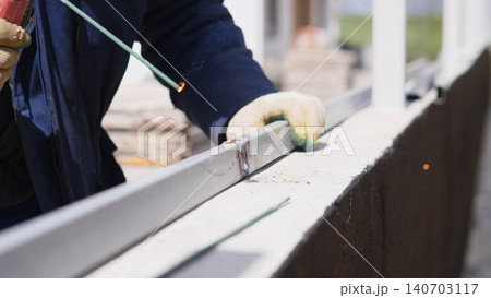 Worker Smoothing Metal Frame With Grinder Wheel, Careful Edge Finishing Near Concrete Barrier, Gloved Hands And Focused Movement Creating Clean Surface And Polished Profile 140703117