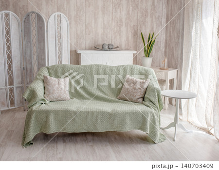 Cozy living room scene featuring a green sofa with decorative pillows, a small white table, and natural light streaming through sheer curtains, creating a warm and inviting atmosphere 140703409