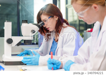 Two female scientists collaborating in a laboratory using a microscope and taking notes on research findings Two female scientists collaborating in a laboratory using a microscope and taking notes on research findings 140704221