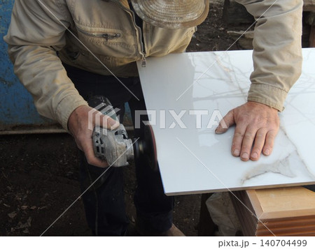The technician holds an angle grinder and grinds at a slight angle to the surface being treated to chamfer or remove chips. The grinding is performed on the edge of large-format white tiles The technician holds an angle grinder and grinds at a slight angle to the surface being treated to chamfer or remove chips. The grinding is performed on the edge of large-format white tiles 140704499