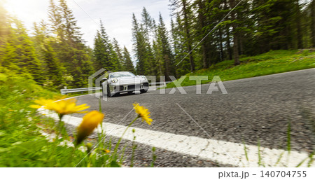 Silver sports car speeding through a winding mountain road surrounded by forest. Dynamic panning shot with motion blur captures performance driving, speed and automotive lifestyle. Silver sports car speeding through a winding mountain road surrounded by forest. Dynamic panning shot with motion blur captures performance driving, speed and automotive lifestyle. 140704755