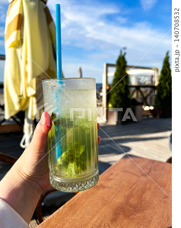 Vertical image of woman hand holding mojito cocktail in transparent glass with ice cubes and straw in summer cafe at beach 140708532