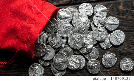 Ancient silver coins spilling from a red cloth pouch onto a dark wooden table. Roman denarii treasure hoard. Numismatics and historical wealth concept 140709125