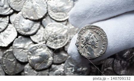 Gloved hand holding an ancient Roman silver coin of Emperor Commodus. Macro close up of a numismatist examining a historical artifact. Pile of old collection in blurred background 140709132