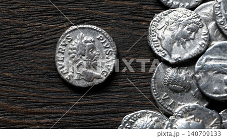 Close up of ancient Roman silver coins on a dark rustic wooden table. Macro view of a denarius with an emperor Septimius Severus. Money, antique and history concept. Close up of ancient Roman silver coins on a dark rustic wooden table. Macro view of a denarius with an emperor Septimius Severus. Money, antique and history concept. 140709133