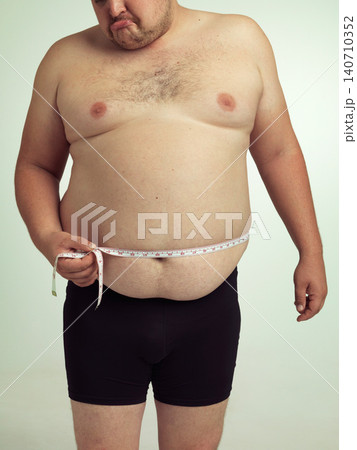 Man, plus size and disappointed on stomach with measurement in studio on white background with tape for weight loss and progress. Sadness, fitness and body with self care to monitor health and shape. Man, plus size and disappointed on stomach with measurement in studio on white background with tape for weight loss and progress. Sadness, fitness and body with self care to monitor health and shape. 140710352