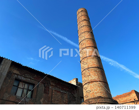 Tall old brick factory chimney rising above abandoned industrial building against blue sky low angle view Tall old brick factory chimney rising above abandoned industrial building against blue sky low angle view 140711399