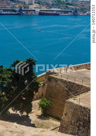 Valletta Grand Harbour Malta vie from limestone bastion walls over blue sea to working industrial port with cargo ships and cranes Valletta Grand Harbour Malta vie from limestone bastion walls over blue sea to working industrial port with cargo ships and cranes 140711553