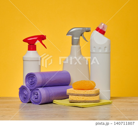 Plastic bottles with household chemicals, rolls of garbage bags, sponges and a napkin stand on a wooden surface Plastic bottles with household chemicals, rolls of garbage bags, sponges and a napkin stand on a wooden surface 140711807