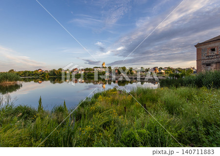 Wide panoramic view of a rural town with a golden dome church reflected in a calm lake. Beautiful summer countryside landscape with historic architecture, green riverbanks, and scenic cloudy sky. 140711883