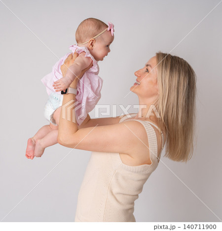 Side view of a happy blonde mother holding her cute baby girl up in the air. Both sharing a joyful, candid moment of connection and love against a clean studio background. A beautiful young mother Side view of a happy blonde mother holding her cute baby girl up in the air. Both sharing a joyful, candid moment of connection and love against a clean studio background. A beautiful young mother 140711900