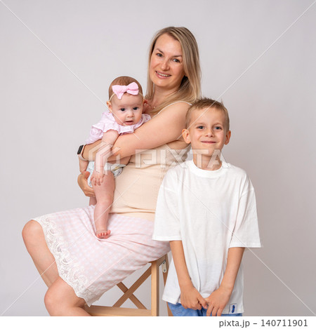 A young blonde mother sits gracefully on a wooden stool holding her baby daughter, while her happy older son stands proudly beside her in a clean, bright studio environment. Beautiful family stock A young blonde mother sits gracefully on a wooden stool holding her baby daughter, while her happy older son stands proudly beside her in a clean, bright studio environment. Beautiful family stock 140711901