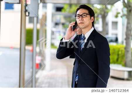 バス停でバスを待ちながら電話するスーツ姿の男性 140712364