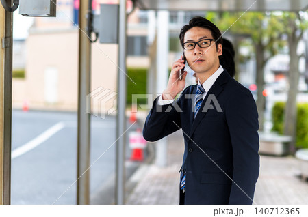 バス停でバスを待ちながら電話するスーツ姿の男性 バス停でバスを待ちながら電話するスーツ姿の男性 140712365