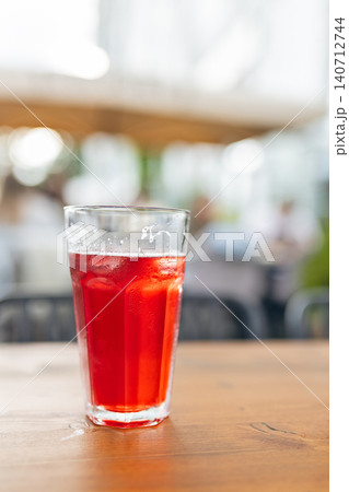 Tall clear glass filled with refreshing red iced beverage or cold brew tea standing on wooden table in outdoor summer cafe with blurry bokeh background 140712744