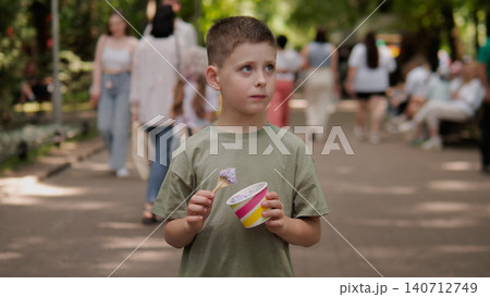 Boy in green shirt holding colorful ice cream cup and spoon while standing on shaded park walkway; soft daylight filtering through trees creates dappled 140712749