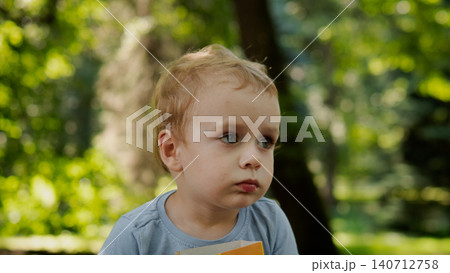 Young toddler boy seated outdoors holding small colorful board book while gazing to side with thoughtful expression, soft sunlight filtering through leafy 140712758