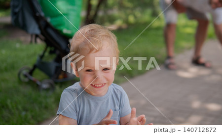 Blond toddler seated on pavement edge clapping hands and smiling at camera during sunny day in green park, soft natural light illuminating curly hair Blond toddler seated on pavement edge clapping hands and smiling at camera during sunny day in green park, soft natural light illuminating curly hair 140712872