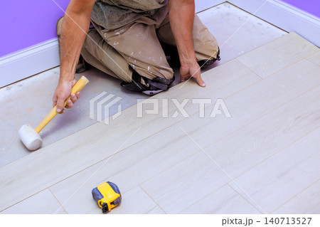 Contractor kneels on laminate floor lays down new planks while using hammer, measuring tape in room renovation 140713527