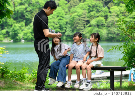 新緑の湖畔のベンチで休憩中に先生から配り物を受け取る女子小学生たち 新緑の湖畔のベンチで休憩中に先生から配り物を受け取る女子小学生たち 140713932