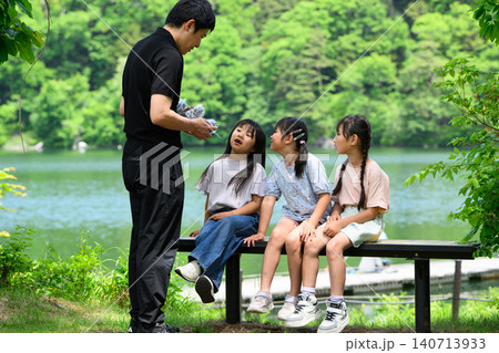 新緑の湖畔のベンチで休憩中に先生から配り物を受け取る女子小学生たち 新緑の湖畔のベンチで休憩中に先生から配り物を受け取る女子小学生たち 140713933