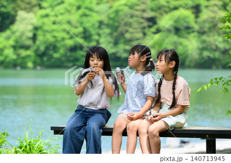 新緑の湖畔のベンチで仲良くおにぎりを食べる女子小学生たち	 140713945