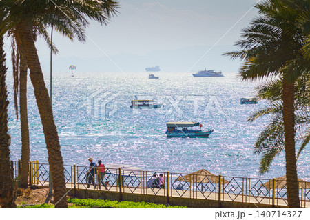 Aqaba, Jordan- Mar 13 2023, a landscape of Red Sea with many boats and park with palm trees and people on the foreground, at daytime, Aqaba, Jordan Aqaba, Jordan- Mar 13 2023, a landscape of Red Sea with many boats and park with palm trees and people on the foreground, at daytime, Aqaba, Jordan 140714327