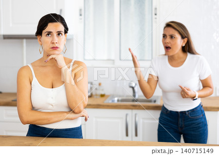 Upset young woman standing in home kitchen while sister reprimanding her 140715149