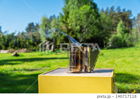 Bee smoker tool emitting smoke on top of a yellow wooden beehive in a sunny green meadow field 140717118