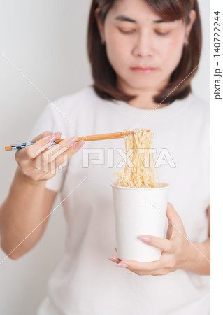 Happy young Asian woman enjoying a delicious cup of hot instant noodles while relaxing at home, Cheerful female eating instant ramen with a smile at apartment. Simple lifestyle and quick meal concept Happy young Asian woman enjoying a delicious cup of hot instant noodles while relaxing at home, Cheerful female eating instant ramen with a smile at apartment. Simple lifestyle and quick meal concept 140722244