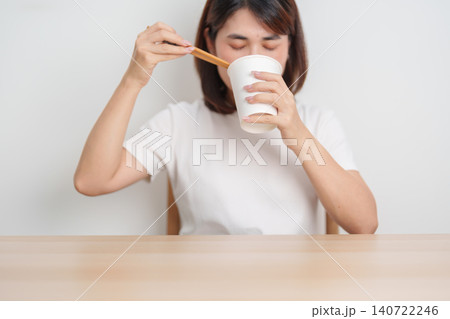 Happy young Asian woman enjoying a delicious cup of hot instant noodles while relaxing at home, Cheerful female eating instant ramen with a smile at apartment. Simple lifestyle and quick meal concept 140722246