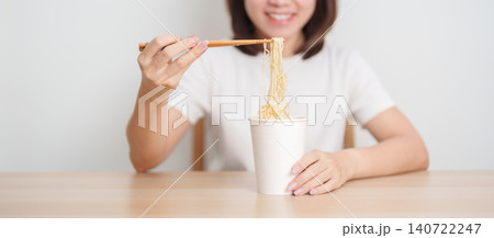 Happy young Asian woman enjoying a delicious cup of hot instant noodles while relaxing at home, Cheerful female eating instant ramen with a smile at apartment. Simple lifestyle and quick meal concept Happy young Asian woman enjoying a delicious cup of hot instant noodles while relaxing at home, Cheerful female eating instant ramen with a smile at apartment. Simple lifestyle and quick meal concept 140722247